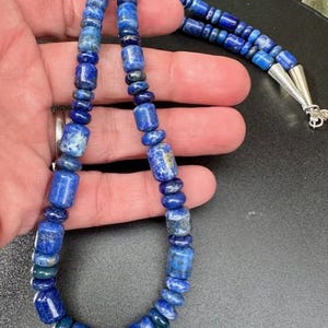 May include: A blue lapis lazuli necklace with a variety of bead shapes, including cylindrical and round beads. The necklace has a silver clasp and is displayed against a dark background.