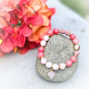 May include: A pink and white beaded bracelet with a gold heart charm. The bracelet is made with natural stones and lava beads. The bracelet is resting on a gray rock.