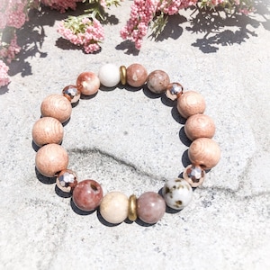 May include: A bracelet made with natural stones and wood beads. The bracelet features a mix of light pink, brown, white, and beige beads. The beads are strung on a gold-colored cord.
