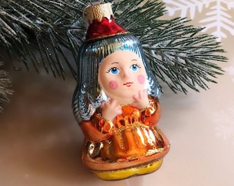 Könnte beinhalten: Ein antiker gläserner Weihnachtsbaumschmuck in Form eines Mädchens, das ein blaues Kleid und einen roten Hut trägt. Sie hat blaue Augen, rosige Wangen und einen süßen Ausdruck.