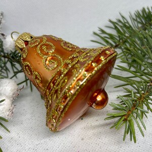May include: A brown glass Christmas bell ornament with gold glitter accents. The bell has a decorative swirl pattern and a small gold loop for hanging. The ornament is set against a backdrop of pine needles and a white surface.
