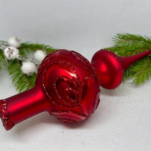 May include: A red glass Christmas tree topper with a swirl design and glitter accents. The topper is shaped like a ball with a pointed top.