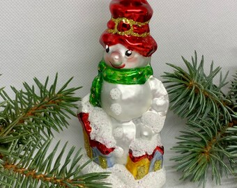May include: A glass snowman Christmas ornament. The snowman wears a red hat with gold trim, a green scarf, and has a smiling face. The ornament is decorated with glitter and sits on a snowy base with miniature houses. A festive holiday decoration.
