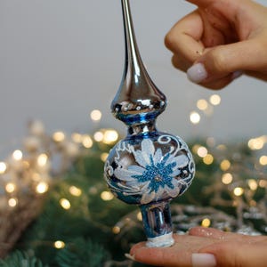 Op de afbeelding: Een blauwe en zilveren glazen kerstboomtopper met een wit bloemmotief. De topper wordt vastgehouden door een hand.