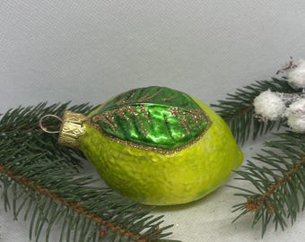 Op de afbeelding: Een kerstversiering in de vorm van een limoen, met een getextureerd, geelgroen lichaam en een groen blad met gouden glitteraccenten. De versiering heeft een goudkleurige metalen dop en een lus om op te hangen. Omgeven door dennentakken.