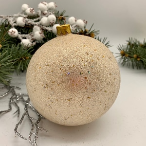 Puede incluir: Una bola de Navidad redonda y color crema con una superficie texturizada y brillante. Tiene una tapa y un lazo de metal dorado para colgar. La vegetación decorativa y las ramas plateadas en el fondo sugieren un tema festivo.