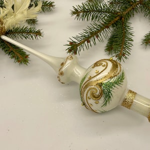 Può includere: Un puntale bianco per albero di Natale con accenti glitterati dorati e dettagli dipinti di verde. Il puntale ha una forma bulbosa con un lungo e sottile stelo. Un piccolo uccello dorato con piume è attaccato alla parte superiore.