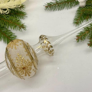 Könnte beinhalten: Ein klarer Weihnachtsbaumspitze aus Glas mit goldenen Glitzerakzenten und einem Schneeflockenmuster. Die Spitze ist von grünen Tannenzweigen umgeben und erzeugt eine festliche Atmosphäre.