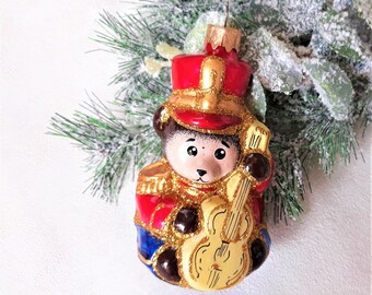 Könnte beinhalten: Ein gläserner Weihnachtsbaumschmuck in Form eines Teddybären, der als Musiker verkleidet ist. Der Bär trägt eine rot-goldene Uniform mit einer blauen Schärpe und hält ein gelbes Cello.