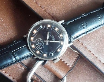 May include: A vintage wristwatch with a black face and a black leather band. The watch face features white and brown markers, silver hands, and a small emblem. The watch is set against a brown leather surface.