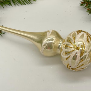 May include: A gold glass Christmas tree topper with white floral designs and gold glitter accents.