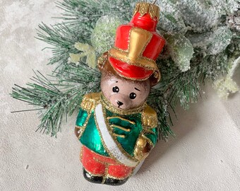 Könnte beinhalten: Ein gläserner Weihnachtsbaumschmuck in Form eines Teddybären, der als Soldat verkleidet ist. Der Bär trägt einen roten Hut mit goldenen Verzierungen, eine grüne Jacke mit goldenen Knöpfen und eine rote Hose. Der Schmuck hat eine goldene und weiße Schärpe.
