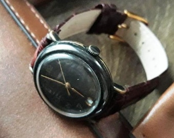 May include: A vintage wristwatch with a dark face and gold hands. The watch has a black case and a burgundy leather band. The watch is resting on a brown leather surface.