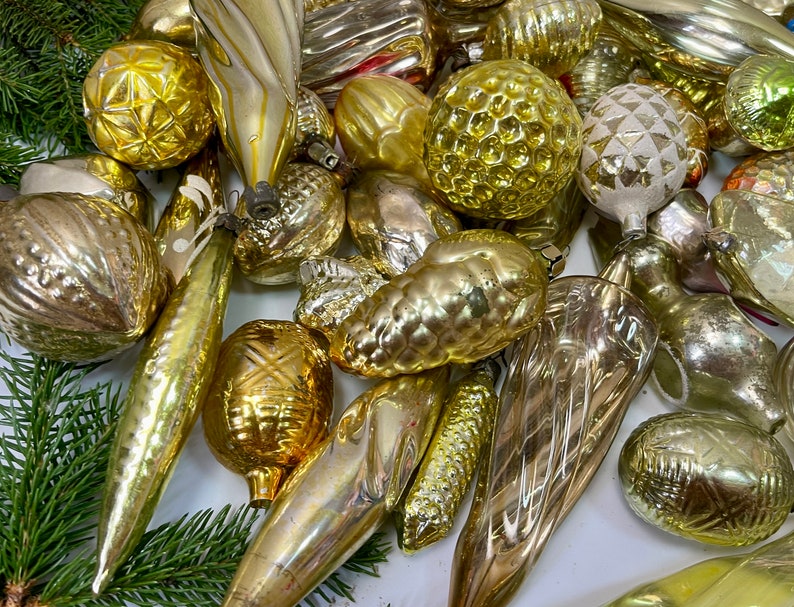 May include: A collection of vintage glass Christmas ornaments in gold and silver tones. The ornaments are various shapes, including balls, teardrops, and cones. Some have intricate patterns and textures.