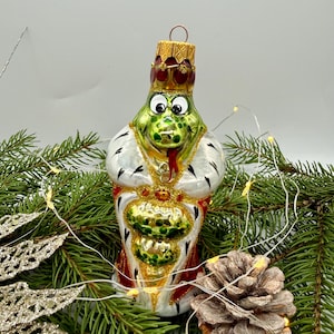 以下が含まれることがあります： 緑色のカエルをかたどったガラス製のクリスマスオーナメント。赤と白のアーミン柄のロイヤルローブと金の王冠を身に着けている。カエルには大きな白い目と赤い舌があり、ローブと王冠には金のグリッターのアクセントが施され、上部には金属製のループが付いている。