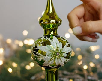Puede incluir: Un adorno para la cima del árbol de Navidad de vidrio verde con un diseño floral blanco y un centro brillante. El adorno tiene una parte superior puntiaguda y una base redonda.