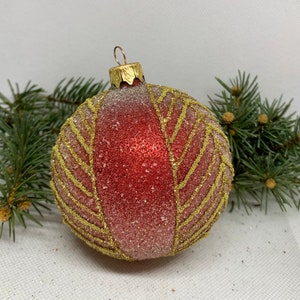 Könnte beinhalten: Ein roter und goldener Weihnachtsschmuck mit strukturierter, glitzernder Oberfläche. Der Schmuck hat ein blattartiges Design aus goldenem Glitzer. Eine goldfarbene Metallkappe und eine Aufhängeschlaufe befinden sich oben. Grüne Kiefernzweige sind im Hintergrund.