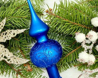 May include: A vibrant blue Christmas tree topper with a decorative, glittery design. The topper is set against a backdrop of green pine needles and festive decorations, creating a holiday theme.