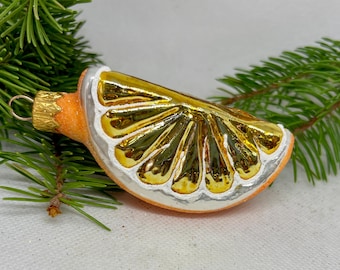 May include: A glass Christmas ornament shaped like a slice of orange. The ornament has a gold interior, white rind, and orange peel. A gold-colored metal loop is attached for hanging. The ornament is displayed with green pine needles.