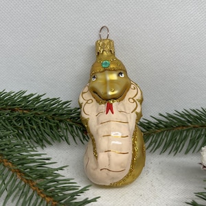 May include: A decorative glass ornament shaped like a cobra snake. The snake is primarily gold and cream-colored, with a red tongue and a green gem on its head. The ornament has a gold cap for hanging and is decorated with glitter.