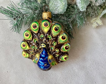May include: A glass peacock Christmas ornament with a brown body, blue chest, and a fan of green and gold feathers. The ornament has a gold metal cap and is hanging near a green and white frosted garland.