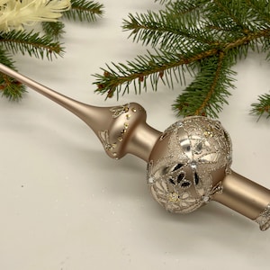 Puede incluir: Un adorno para la parte superior del árbol de Navidad de color champán con un tallo largo y delgado y un globo decorativo. El globo está adornado con intrincados filigranas plateados y acentos brillantes. Ramas de pino verdes en el fondo.
