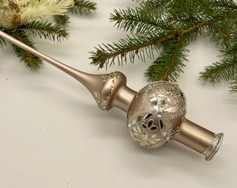 May include: A champagne-colored Christmas tree topper with a long, slender stem and a decorative globe. The globe is adorned with intricate silver filigree and sparkling accents. Green pine branches are in the background.