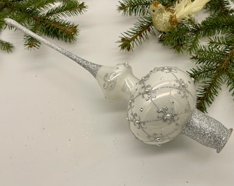 Puede incluir: Adorno para la cima del árbol de Navidad de vidrio blanco con acabado de purpurina plateada y un delicado diseño plateado. El adorno tiene una punta puntiaguda y una base redonda.