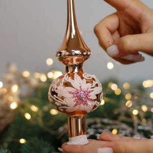 May include: A rose gold Christmas tree topper with a white and pink floral design. The topper has a pointed top and a round base.