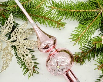 May include: A pink Christmas tree topper with a silver glitter base and tip. The topper has a bulbous shape with a floral design and is surrounded by green pine needles and a gold glittery decorative element.