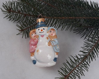 May include: A white ceramic Christmas ornament featuring a snowman with two children holding onto him. The snowman is wearing a blue hat and has a red heart on his chest. The children are wearing pink and blue clothing.
