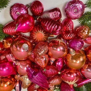 May include: A collection of vintage glass Christmas ornaments in shades of pink and peach. The ornaments are all different shapes and sizes, and some have intricate designs.