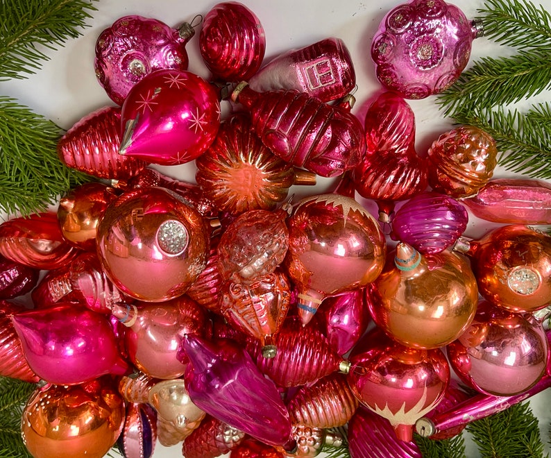 May include: A collection of vintage glass Christmas ornaments in shades of pink and peach. The ornaments are all different shapes and sizes, and some have intricate designs.