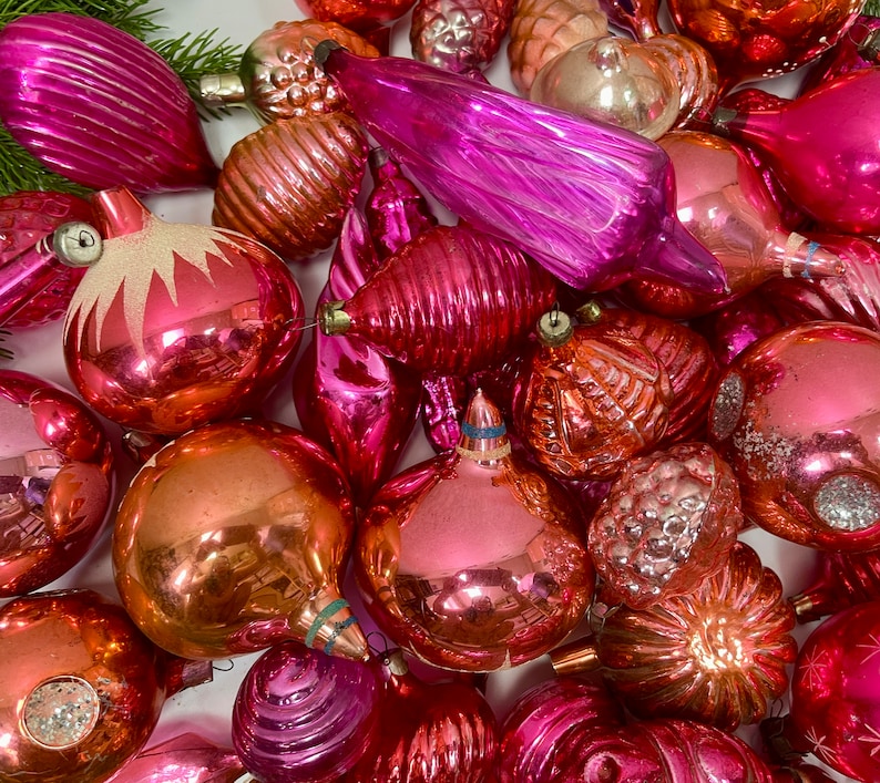May include: A collection of vintage glass ornaments in various shades of pink and red. The ornaments are all different shapes and sizes, and some have a textured or iridescent finish.