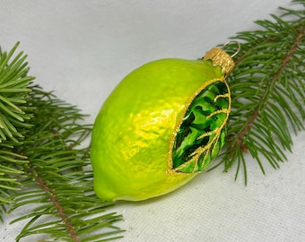Op de afbeelding: Een kerstversiering in de vorm van een limoen met een getextureerd, heldergroen oppervlak. Aan de zijkant is een gedetailleerd bladerontwerp met gouden glitteraccenten zichtbaar. De versiering heeft een goudkleurige metalen dop en ligt op dennennaalden.