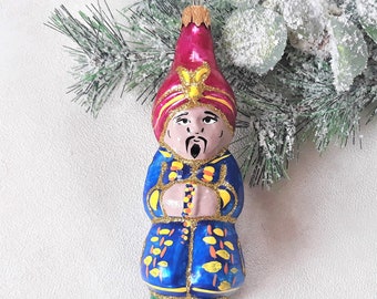 May include: A glass Christmas ornament shaped like a person wearing a blue robe, a red turban, and a gold trim. The ornament has a painted face with a mustache and a beard.
