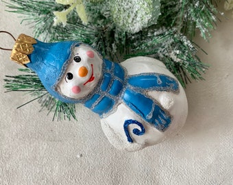 May include: A snowman Christmas ornament with a blue hat and scarf, a carrot nose, and a smiling face. The ornament has a gold-colored top and is decorated with glitter. The snowman is white with blue accents.