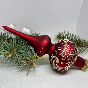 May include: A red glass Christmas tree topper with white and gold painted floral designs. The topper has a pointed top and a bulbous bottom.