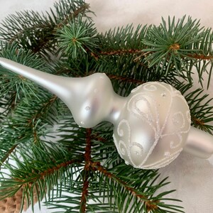 Op de afbeelding: Een witte glazen kerstboomtopper met een delicaat, wervelend ontwerp. De topper heeft een matte afwerking en een puntige punt.
