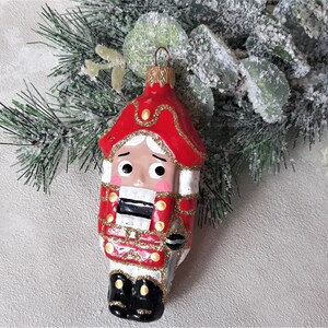 May include: A red and white glass Christmas ornament shaped like a nutcracker soldier. The ornament has a gold trim and a black hat.