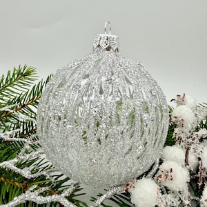 Könnte beinhalten: Ein silberner Weihnachtsbaumschmuck mit strukturierter, glitzernder Oberfläche. Der runde Schmuck wird auf einem Bett aus immergrünen Zweigen und weißen Beeren präsentiert und schafft eine festliche Urlaubsszene. Der Schmuck hat eine Metallkappe und eine Schlaufe zum Aufhängen.