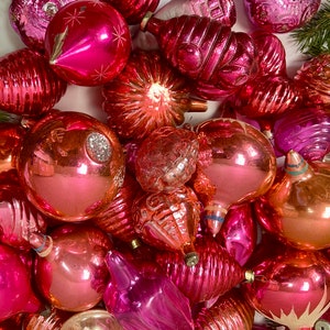 May include: A collection of vintage pink and peach glass Christmas ornaments. The ornaments are various shapes and sizes, including round, teardrop, and star shapes. Some of the ornaments have a shiny finish, while others have a matte finish.