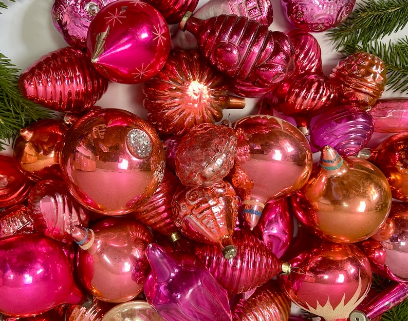 May include: A collection of vintage pink and peach glass Christmas ornaments. The ornaments are various shapes and sizes, including round, teardrop, and star shapes. Some of the ornaments have a shiny finish, while others have a matte finish.