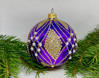May include: A purple Christmas ornament with gold glitter accents and faux pearl embellishments. The ornament features a diamond pattern and is surrounded by green pine branches. The ornament has a gold metal cap.