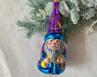 Könnte beinhalten: Ein gläserner Weihnachtsbaumschmuck in Form eines Zauberers mit einem lila Hut und einem blauen Gewand. Der Zauberer hat einen weißen Bart und einen gelben Stern auf seinem Hut.