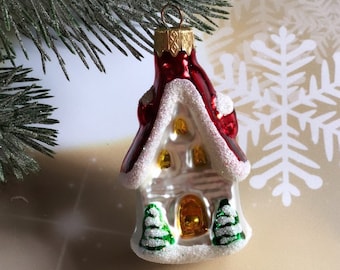 Könnte beinhalten: Ein gläserner Weihnachtsbaumschmuck in Form eines weißen Hauses mit rotem Dach und schneebedeckten Details. Das Haus hat zwei kleine grüne Bäume davor und ein gelbes Fenster.
