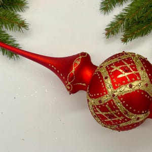 May include: A red and gold Christmas tree topper, featuring a long stem and a patterned globe. The globe is adorned with gold detailing and a grid pattern. Festive decoration for the holiday season.
