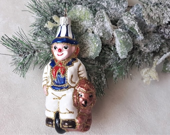 Könnte beinhalten: Ein antiker gläserner Weihnachtsbaumschmuck, der einen Clown in einer blau-weiß gestreiften Mütze zeigt, der einen braunen Hund hält. Der Clown trägt einen weißen Anzug mit goldenen Verzierungen und einen roten Schal.