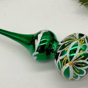 May include: Two green glass Christmas tree toppers with white and gold floral designs. One topper has a long, pointed tip with a glittery white end. The other has a rounded shape with a glittery white base. Festive holiday decor.