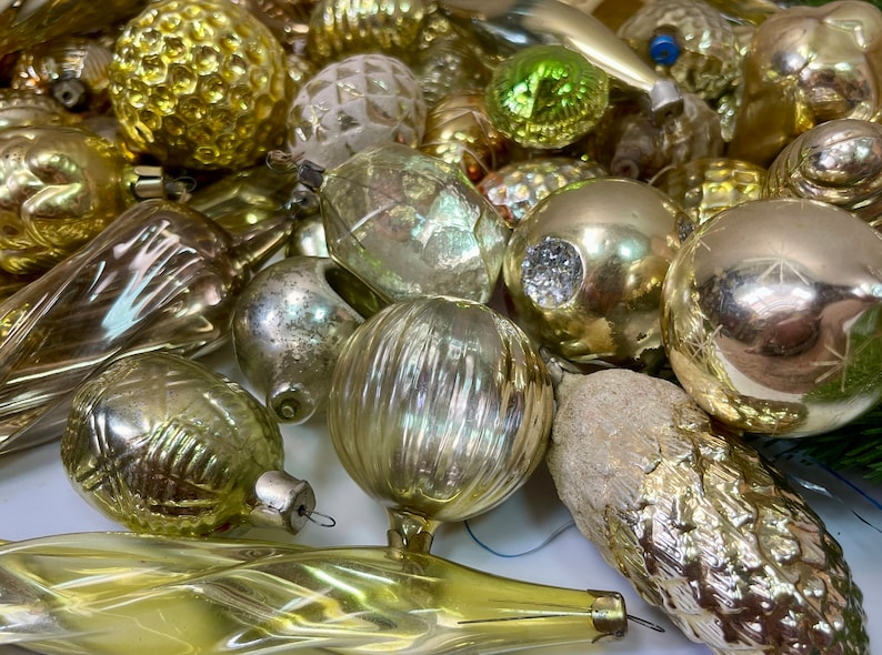 May include: A collection of vintage glass Christmas ornaments in various shapes and sizes, including balls, teardrops, and cones. The ornaments are mostly gold and silver with some clear glass ornaments.